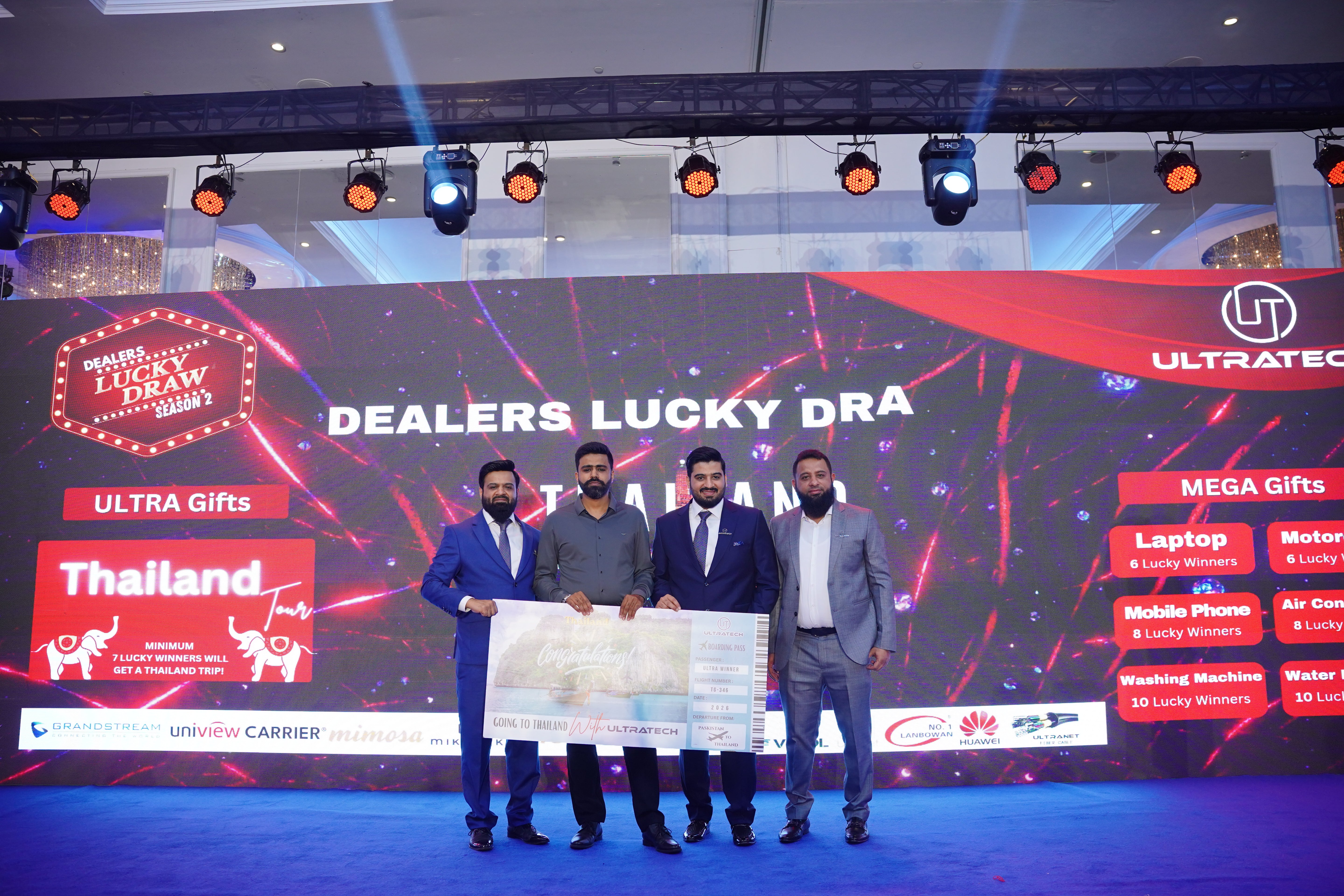 Group of men holding a large check on a stage with a 'Dealers Lucky Draw 2026' banner in the background.